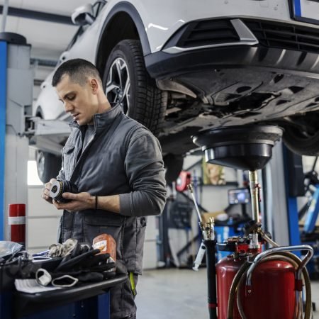 A mechanic's shop worker is cleaning engine parts and fixing it while standing i garage with car on a lift in a background.